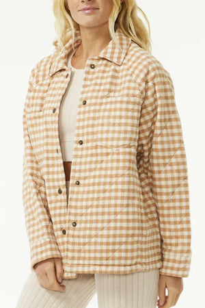 A person wears the Rip Curl Premium Quilted Check Jacket in peach and white gingham with snap buttons and long sleeves, made from yarn dyed fabric. They pair it with a light beige top and cream pants against a plain white background.