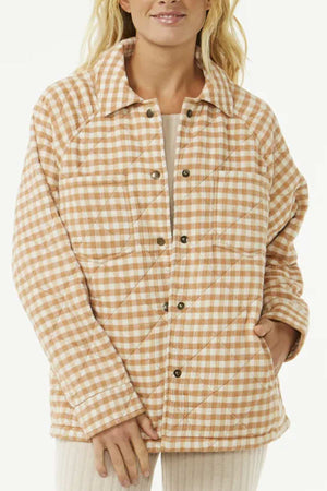 A woman with long blonde hair stands against a plain light background, wearing the Rip Curl Premium Quilted Check Jacket in beige and white, featuring snap buttons, a collar, side pockets, and soft Cotton Double Weave Gauze.