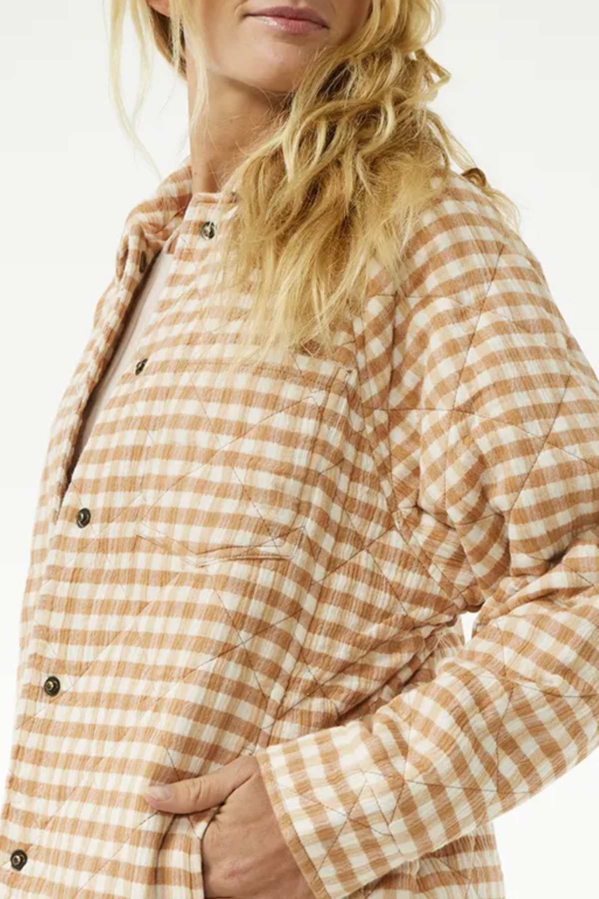 A person with long, wavy blonde hair is wearing the Rip Curl Premium Quilted Check Jacket in orange and white plaid with black buttons. Their left hand is tucked into a side pocket. The background is a plain light color.