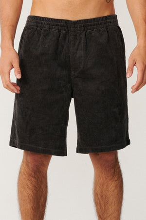 A person wearing Rip Curl Premium Surf Cord 19" Volley black corduroy shorts stands against a plain light background, with only their lower torso and upper legs visible.