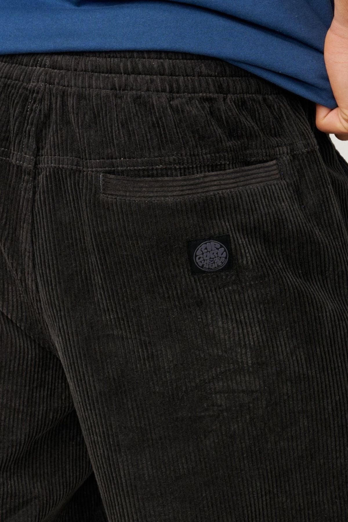 Close-up of a person wearing the Rip Curl Premium Surf Cord 19" Volley in black with a blue shirt. The shorts feature a small back pocket accented by a circular logo above it. Inspired by classic surf volley designs, they combine style and practicality. The person's hand rests partially visible at their side.