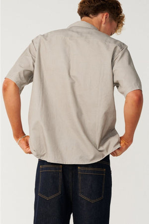 A person is shown from the back, wearing a light gray Rip Curl Premium Surf Cord Short Sleeve Shirt paired with dark blue jeans. The individual has short, curly hair and stands against a plain light backdrop, creating an effortlessly casual look.