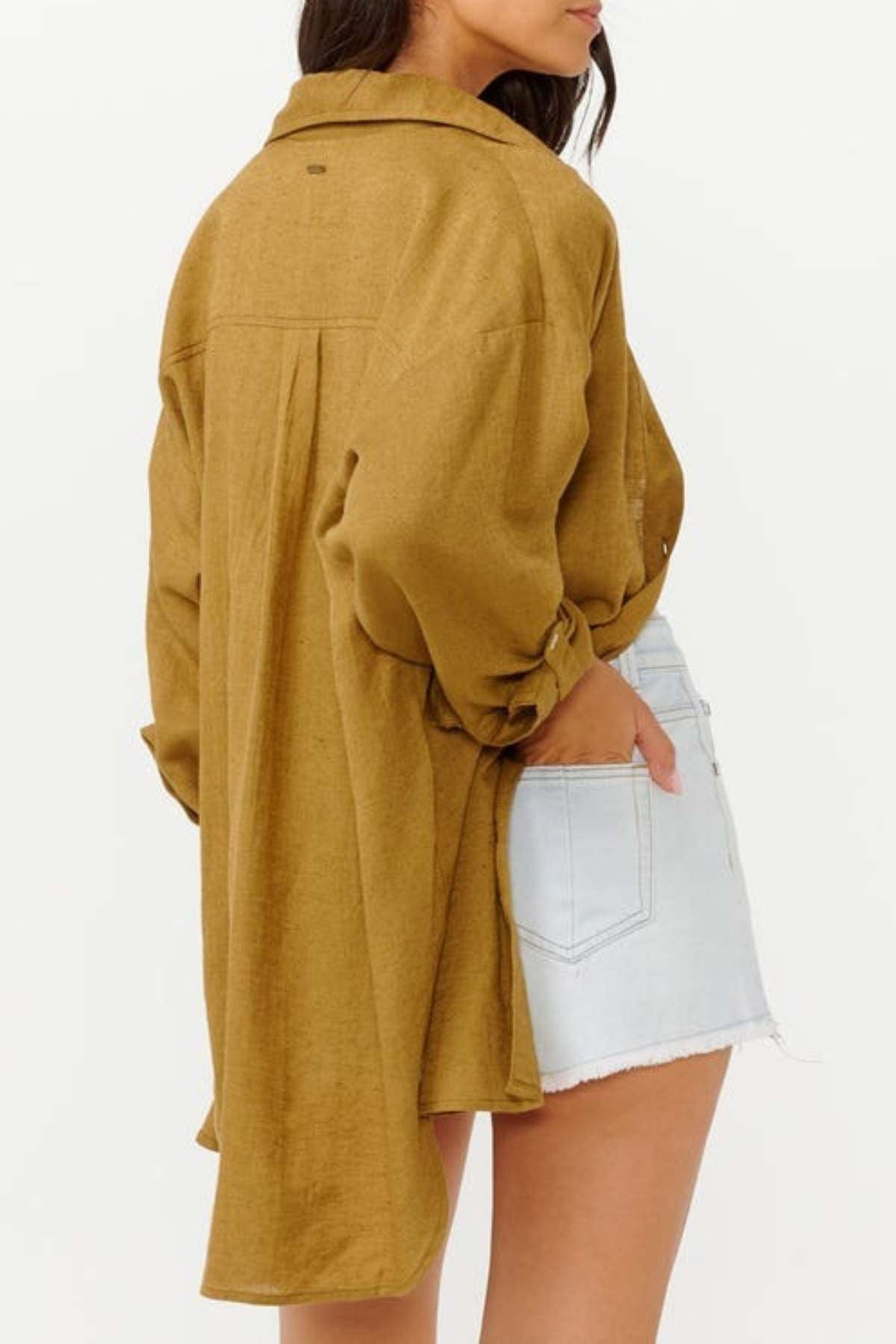 A person stands with their back to the camera, exuding a laid-back surf style in a relaxed fit, mustard-colored Rip Curl Premium Surf Holiday Shirt and light denim shorts. Their left hand is casually tucked into the shorts pocket against a plain white backdrop.