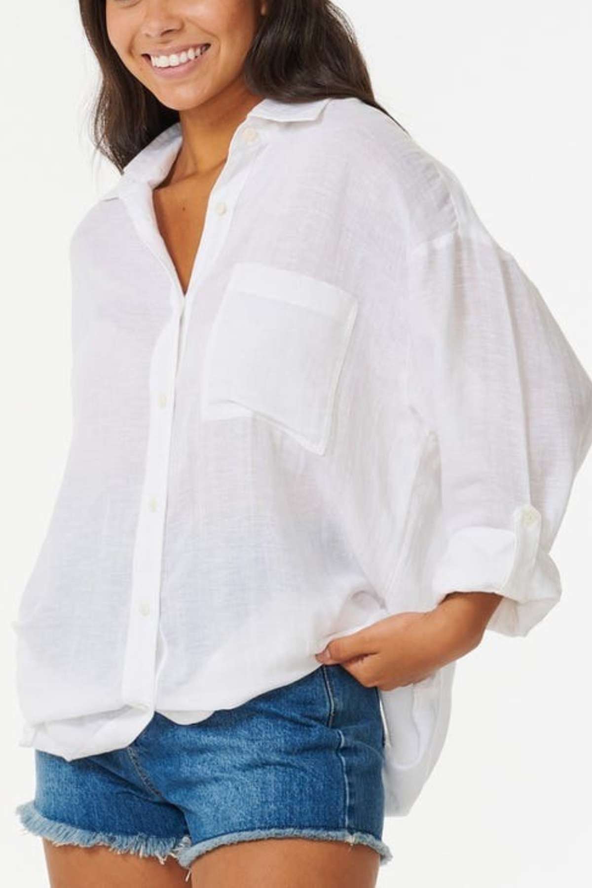 A person wearing a Rip Curl Premium Surf Holiday Shirt with rolled sleeves and denim shorts stands against a plain background, smiling slightly, as if ready for a surf holiday.