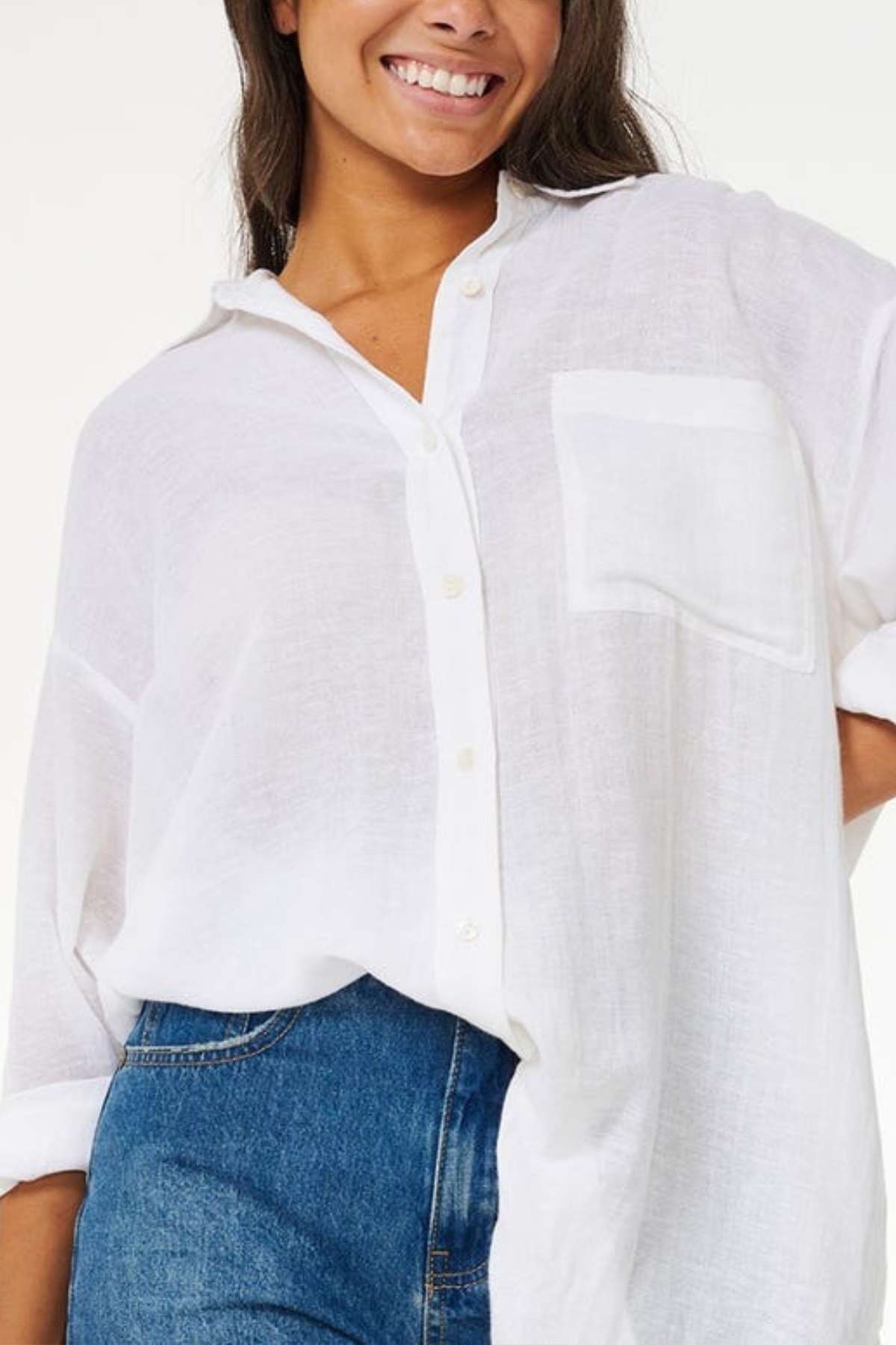 A person is smiling while wearing the Rip Curl Premium Surf Holiday Shirt, a relaxed fit white button-up with rolled-up sleeves and a chest pocket, paired with blue jeans. The background is plain and light-colored.