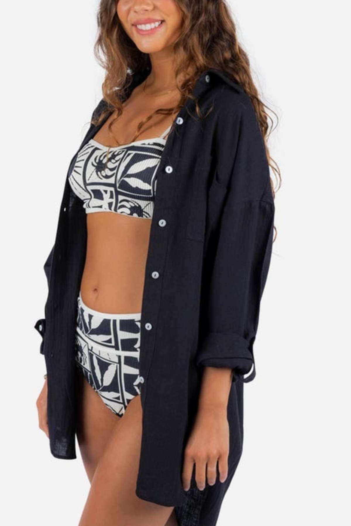 A person wearing a black and white patterned bikini paired with the Rip Curl Premium Surf Holiday Shirt with sleeves rolled up. The individual, who has long, wavy hair and a joyful smile against a plain background, sports an outfit featuring subtle metal logo trim.