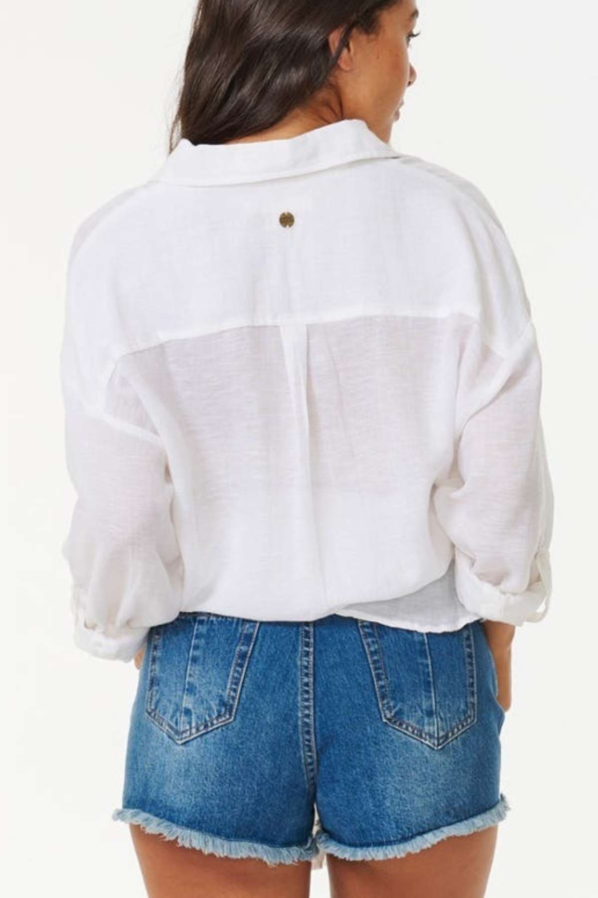A woman with long dark hair is wearing the Rip Curl Premium Surf Holiday Shirt paired with blue denim shorts. She stands with her back to the camera against a plain white background, capturing a look of effortless style and comfort.