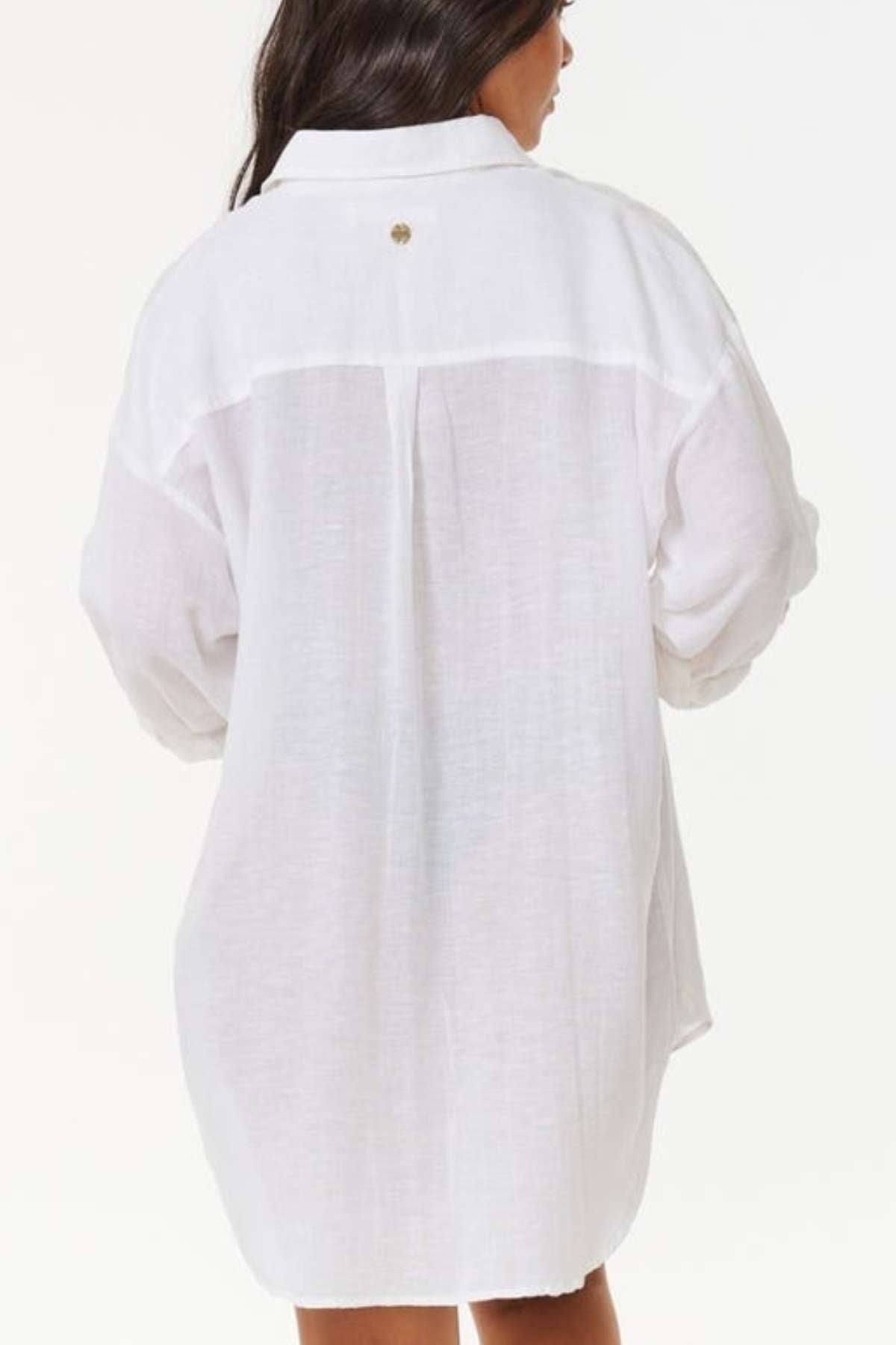 A person with long dark hair is wearing the Rip Curl Premium Surf Holiday Shirt, an oversized white linen piece that exudes an effortless surf holiday vibe. The back is adorned with a small button detail, and the plain, bright background perfectly highlights the shirt's relaxed fit.