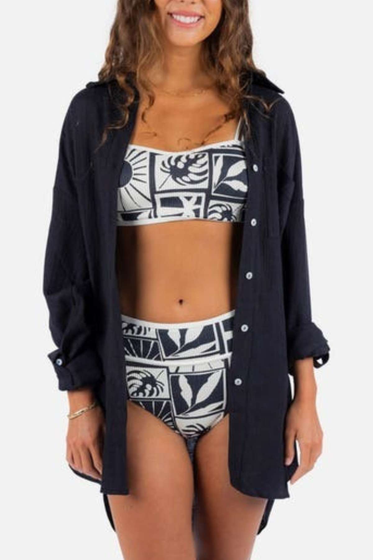 A woman wearing a patterned two-piece swimsuit and an open, relaxed fit shirt is smiling against a plain background. Her wavy hair complements the bracelets on her wrist. The ensemble, featuring the Rip Curl Premium Surf Holiday Shirt, captures a breezy vibe effortlessly.