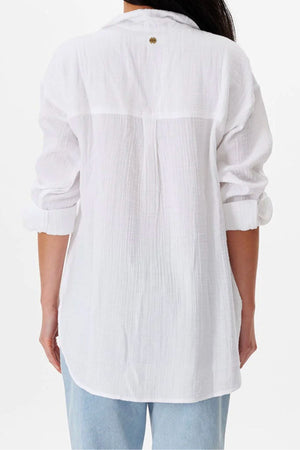 A person faces away, wearing the Rip Curl Premium Surf Long Sleeve Button Up Shirt in a relaxed fit with rolled-up sleeves, paired with light blue jeans. The white surf-style shirt extends below the hips for casual comfort.