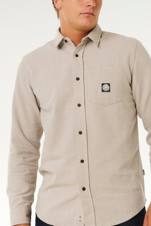 A man wearing a light beige Rip Curl Premium Surf Flannel with a black and white patch on the chest pocket, standing against a plain white background.