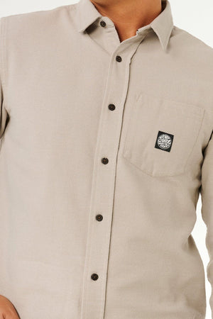 A person wears the Rip Curl Premium Surf Flannel, a light beige long sleeve shirt with dark buttons and a chest pocket featuring a small black and white circular patch, perfect for capturing a relaxed coastal vibe. The person's face is not visible.
