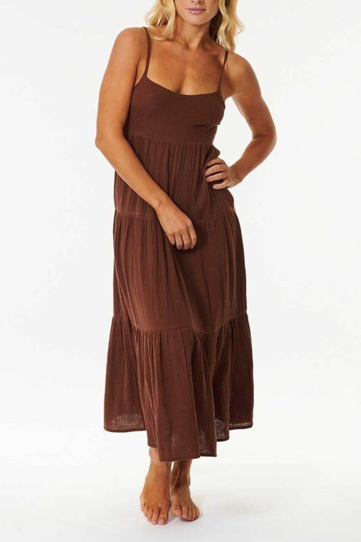 A woman wears the Rip Curl Premium Surf Midi Dress—a sleeveless, brown tiered midi made from cotton double weave gauze with spaghetti straps—standing barefoot against a white background. Perfect for summer occasions.