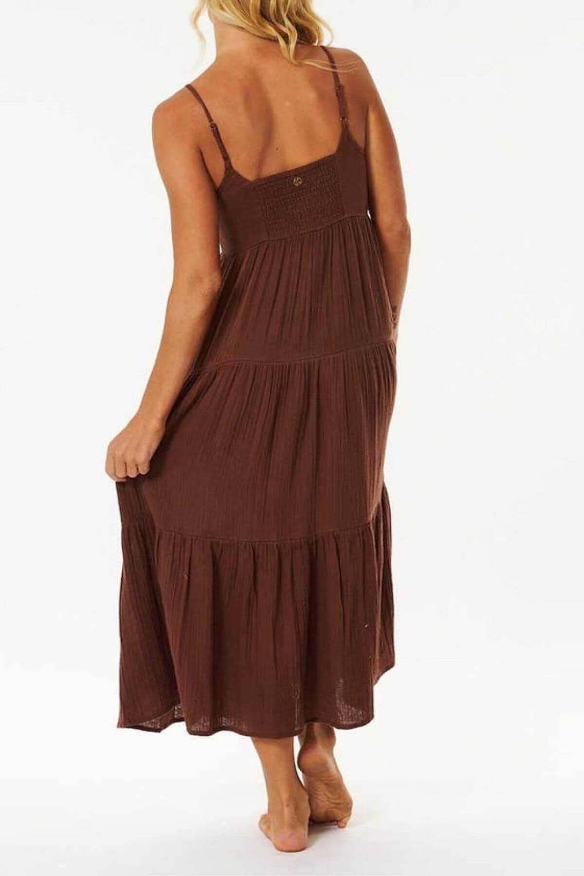 A blonde woman stands barefoot with her back to the camera, holding one side of the Rip Curl Premium Surf Midi Dress made from cotton double weave gauze against a plain white background—an ideal look for summer occasions.