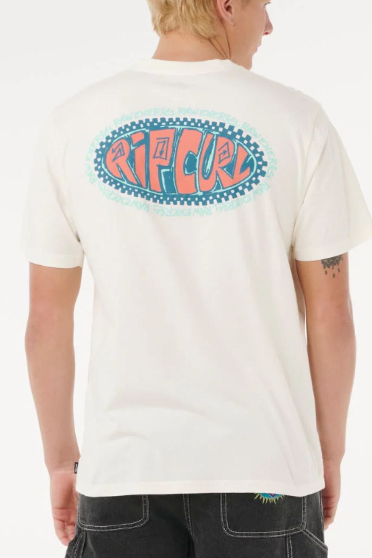 A person with short blond hair is wearing a Rip Curl Raw Energy De Oval Tee, a white 100% cotton short-sleeve shirt with a large blue, teal, and orange "RIP CURL" logo on the back, paired with dark shorts.