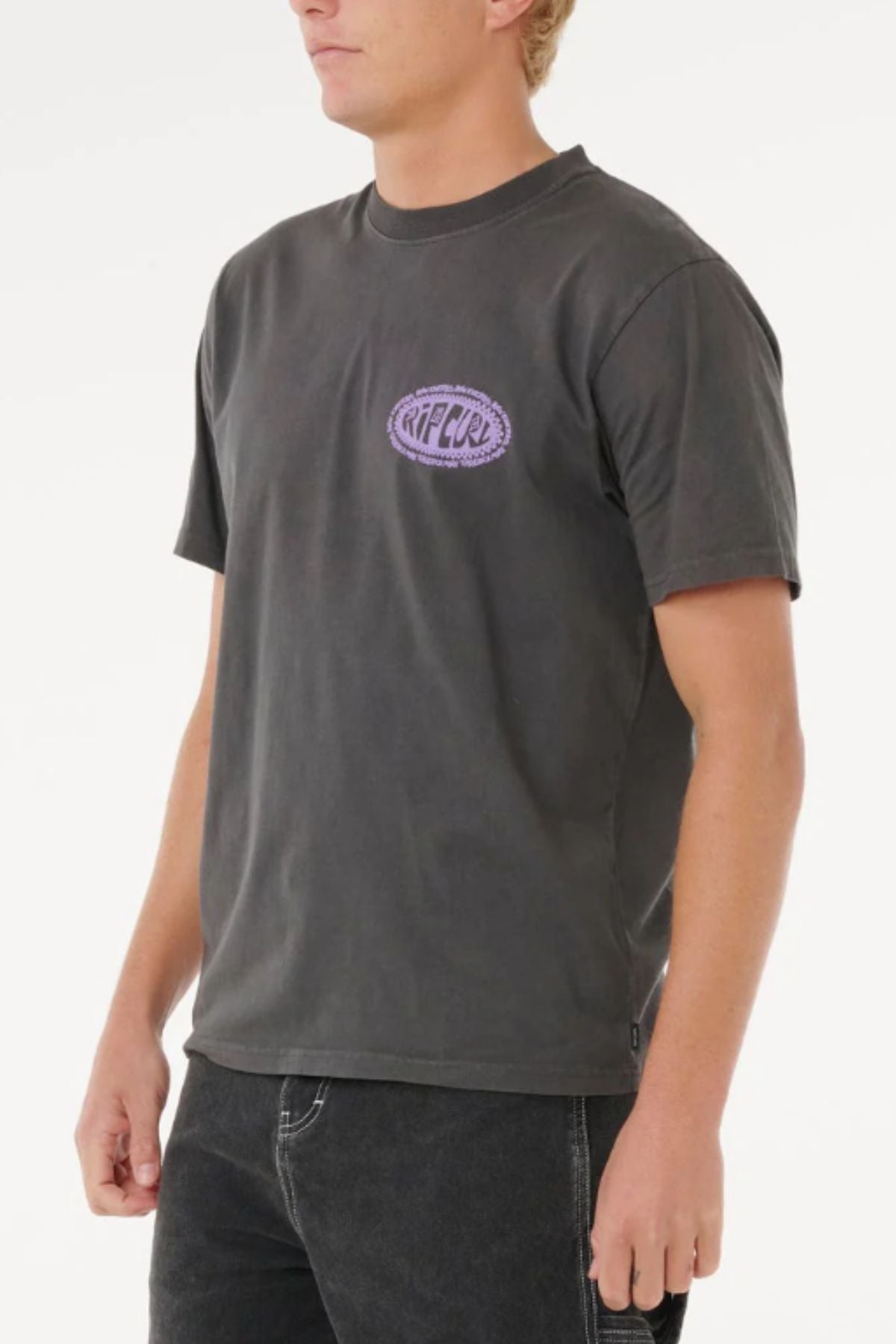 A person wearing a faded black Rip Curl Raw Energy De Oval Tee, a men's cotton short-sleeve shirt with a small purple Rip Curl logo on the left chest, paired with black jeans and standing against a plain white background.