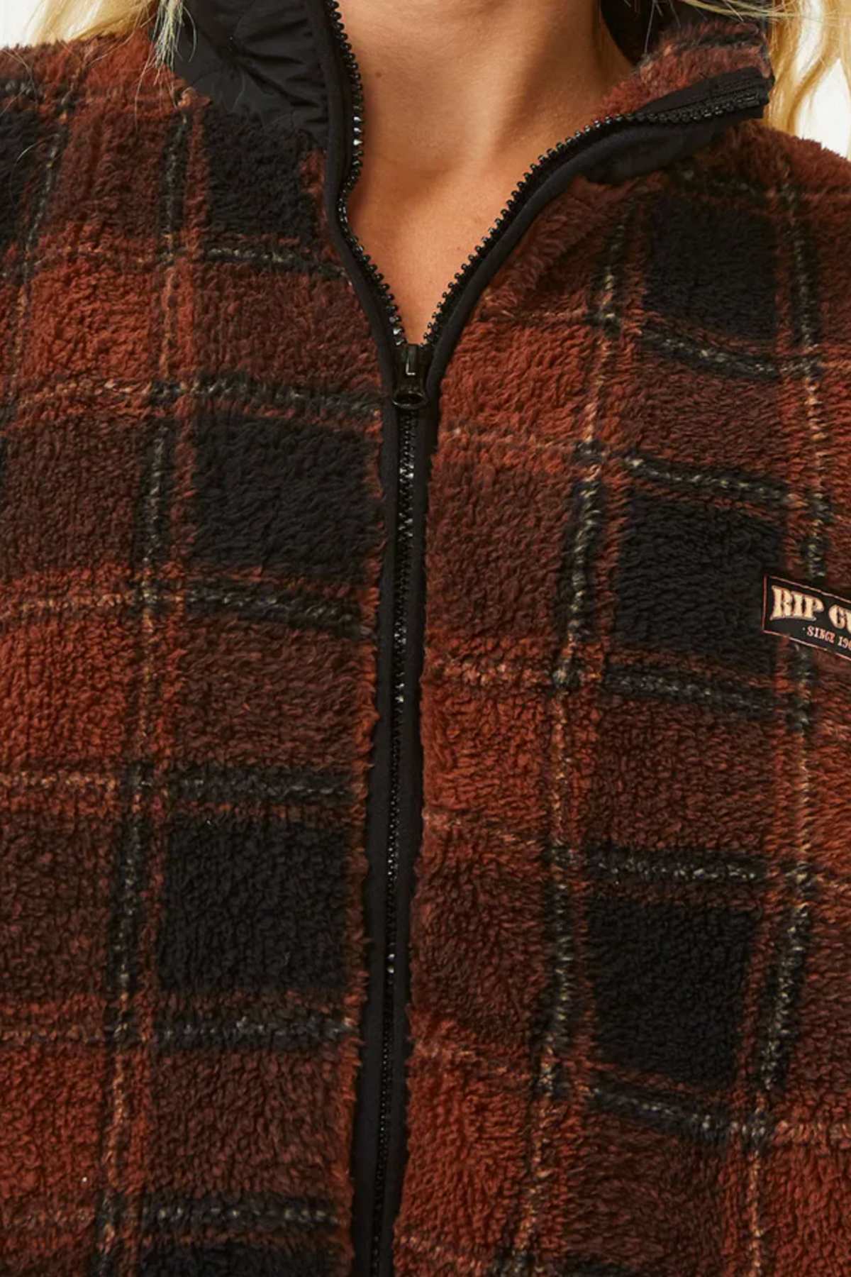 Close-up of a person wearing the Rip Curl Sea Of Dreams Check Fleece, a stylish plaid jacket in brown, black, and red with a zip-up front and small "RIP CURL" logo patch on the left chest.