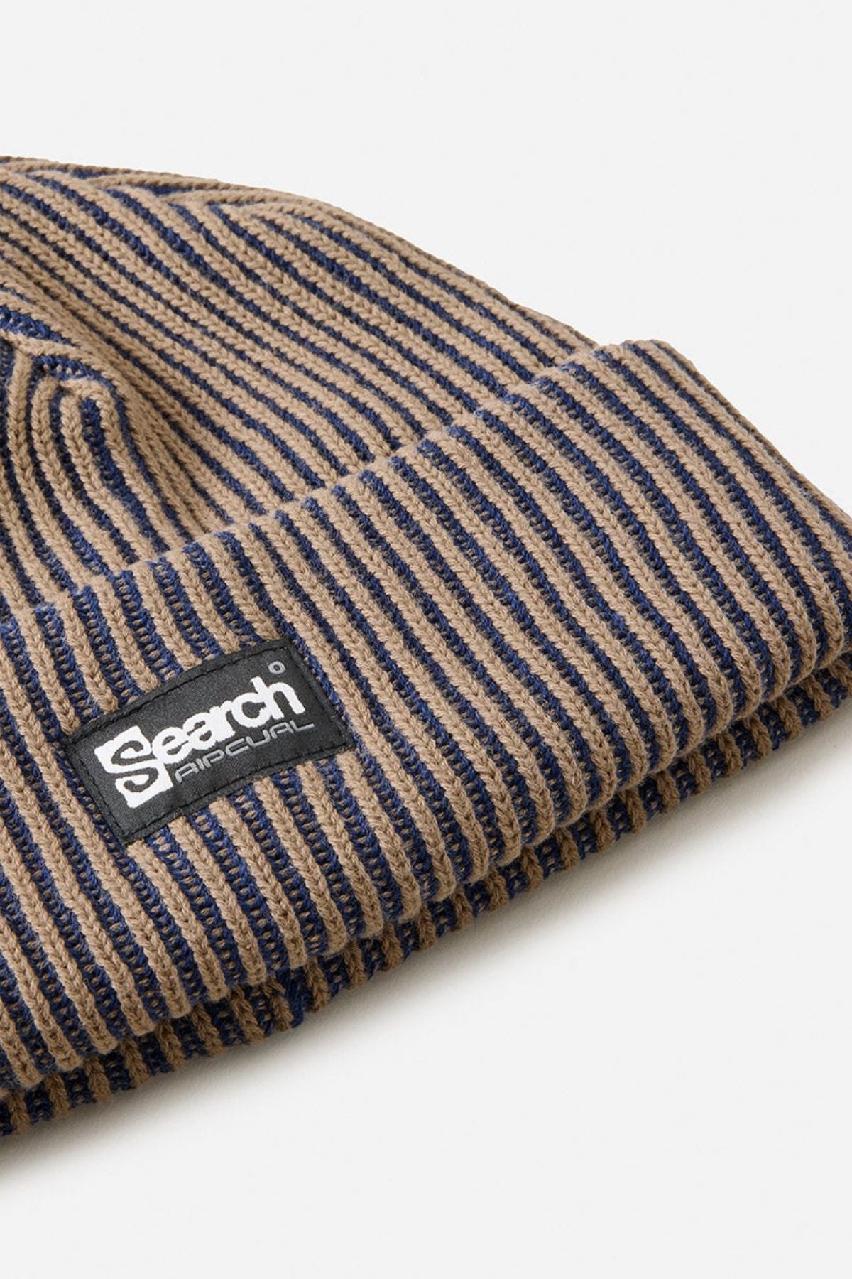 A close-up of the Rip Curl Search Chunky Reg Beanie, a ribbed knit winter hat with brown and navy stripes, featuring a black rectangular patch reading “Search.” This cozy cotton beanie is perfect for chilly days.