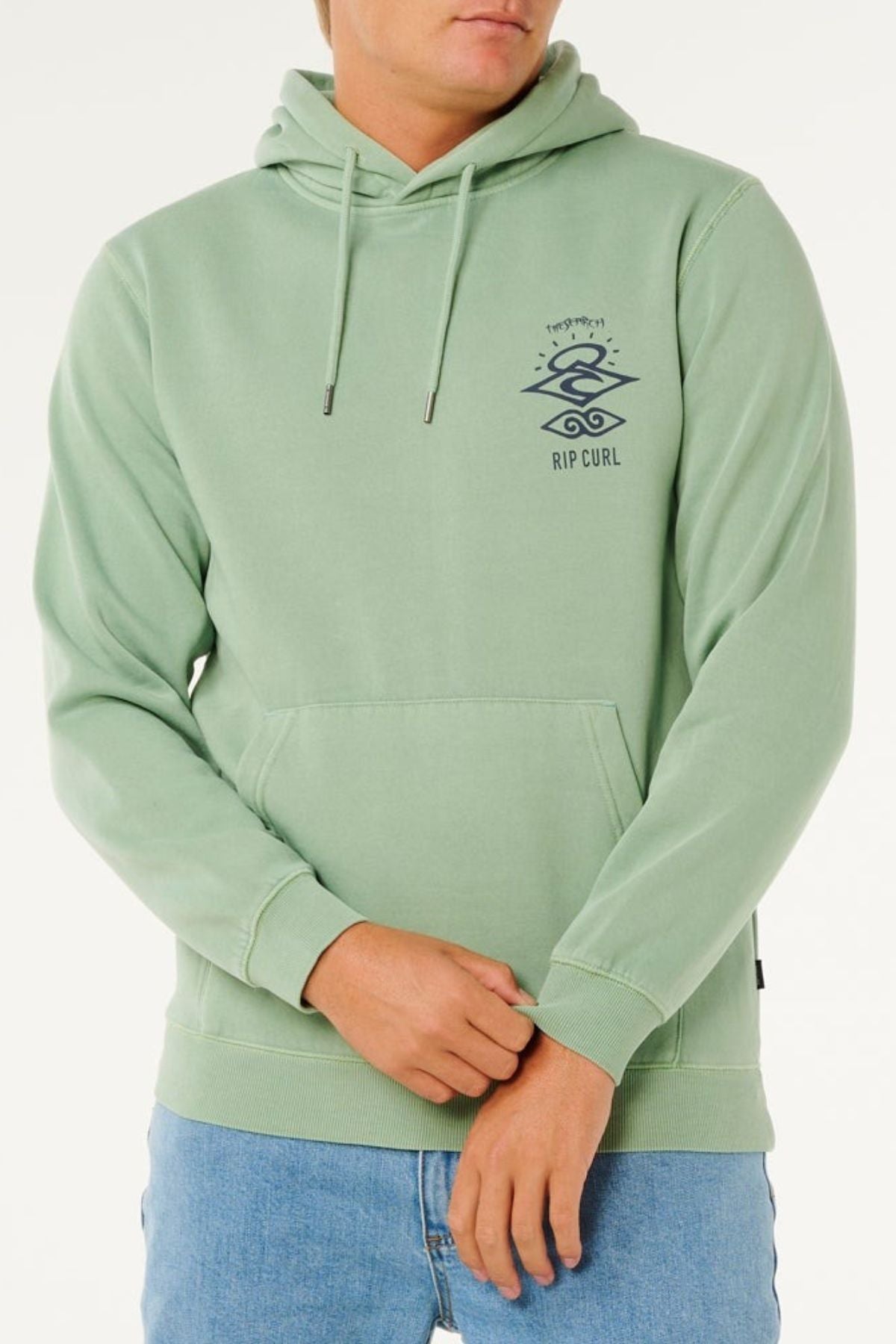 A person wears a light green Rip Curl Search Icon Hood made from organic cotton, featuring a small logo on the left chest, paired with light blue jeans, standing against a plain light background.