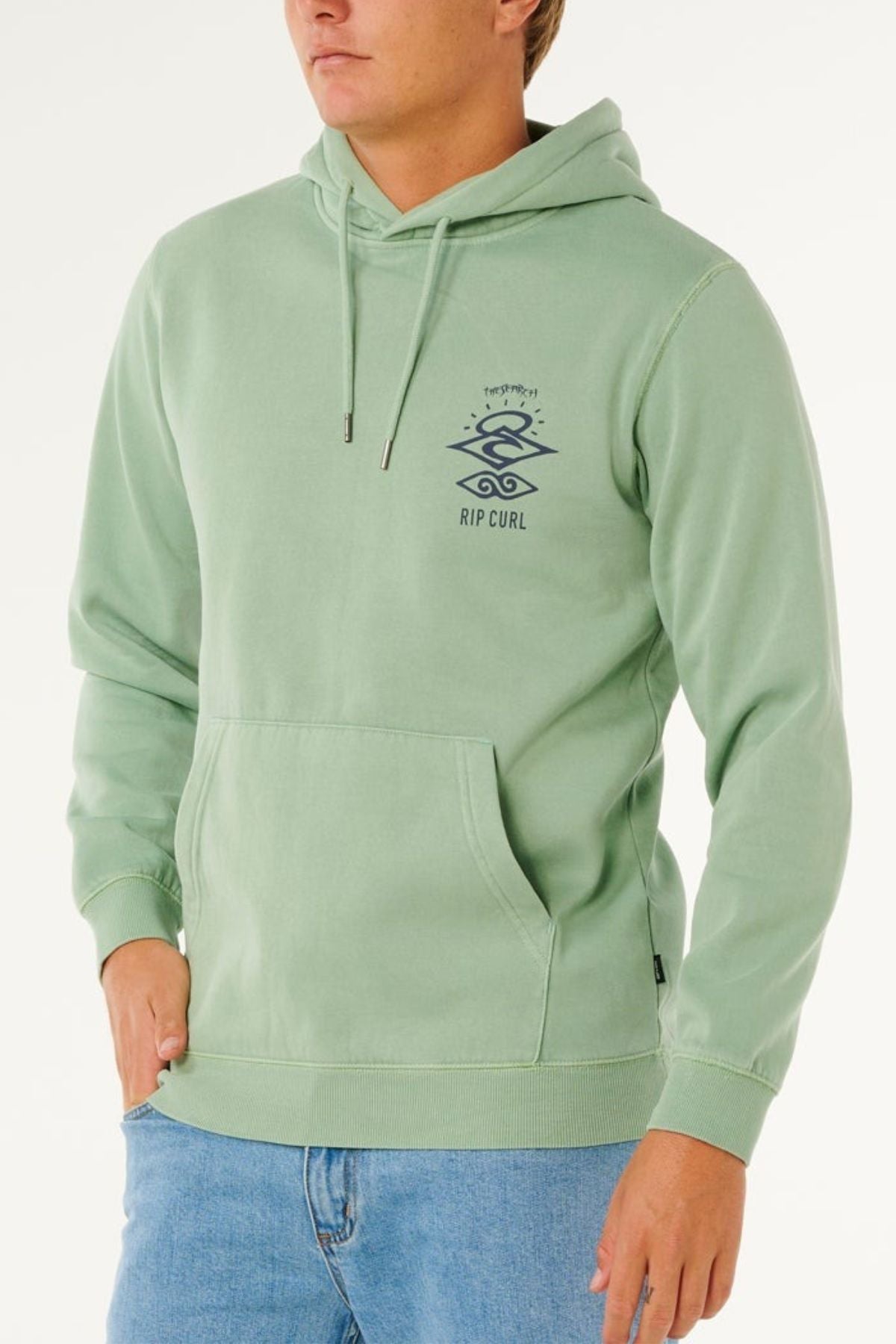A person wears a light green Rip Curl Search Icon Hood made from organic cotton, featuring a front pocket and small dark logo on the left chest, paired with blue jeans, standing against a plain light background.