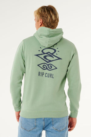 A blonde-haired person faces away from the camera, wearing the Rip Curl Search Icon Hood in light green with a dark blue abstract design and "RIP CURL" on the back, paired with light blue jeans.