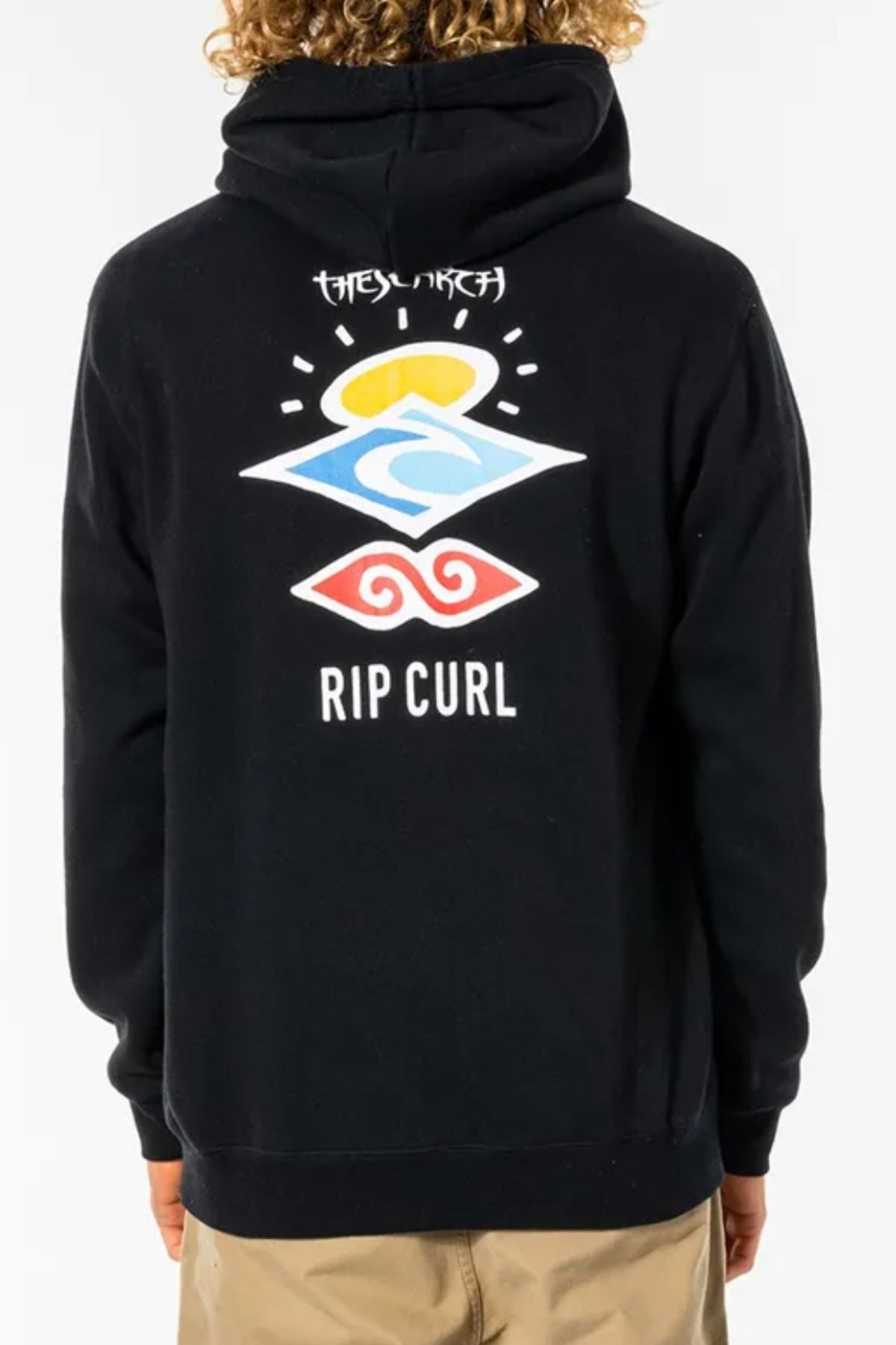 A person with curly hair, facing away from the camera, wears a black Rip Curl Search Icon Hood with colorful graphic symbols and “RIP CURL” text on the back, paired with tan pants.