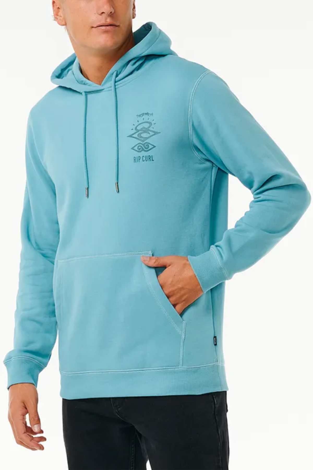 A person wearing a Rip Curl Search Icon Hood in light blue, featuring "Rip Curl" and a logo printed on the left chest. The cozy fleece lining and Organic Brushed Cotton provide exceptional comfort. With one hand tucked into the front pocket of the hoodie, the individual pairs it with black pants, standing against a plain white background.