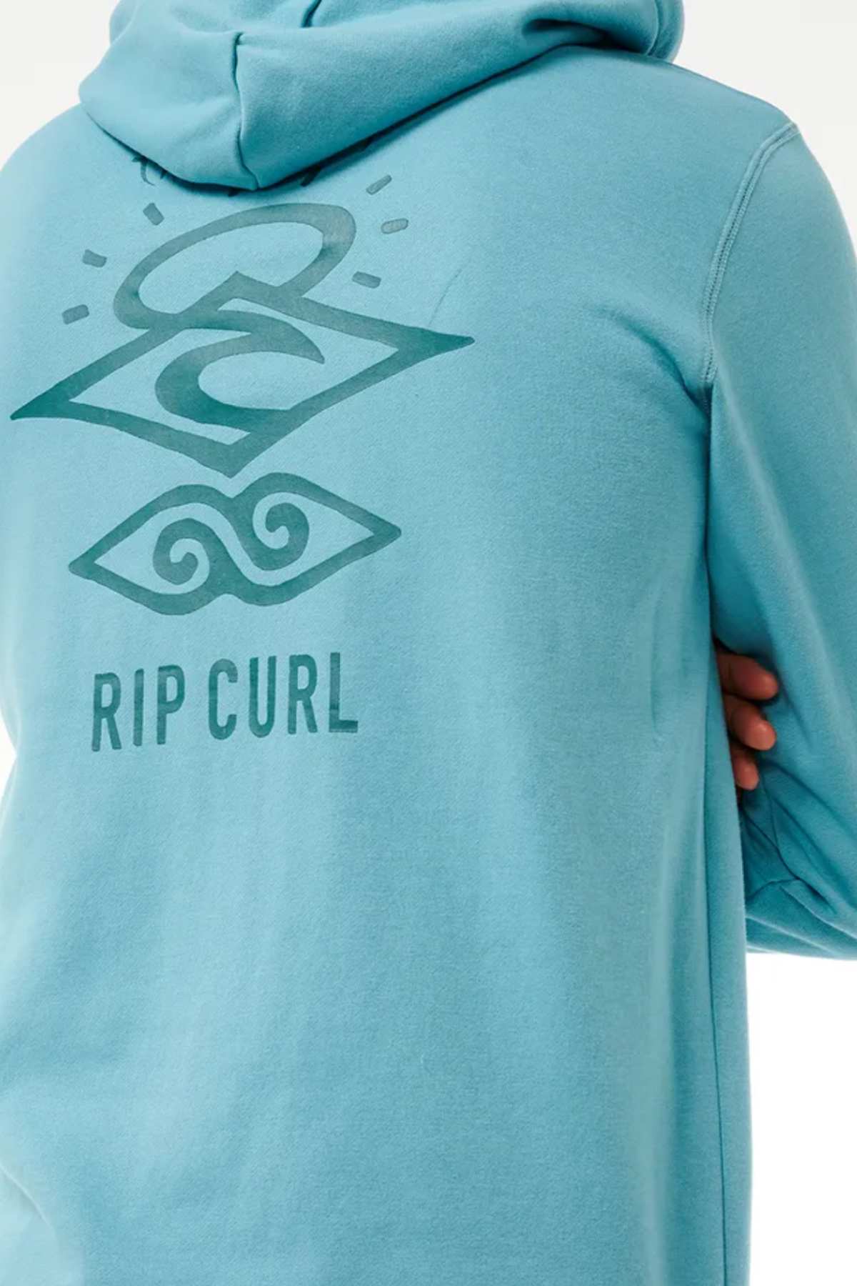 A person wearing a teal Rip Curl Search Icon Hood with the hood up. The back of the Organic Brushed Cotton hoodie features a graphic design in darker teal, including the words "RIP CURL" and two stylized symbols, one resembling a wave and the other resembling a sun and ocean.