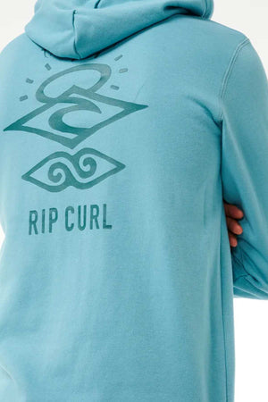 A person wearing a teal Rip Curl Search Icon Hood with the hood up. The back of the Organic Brushed Cotton hoodie features a graphic design in darker teal, including the words "RIP CURL" and two stylized symbols, one resembling a wave and the other resembling a sun and ocean.