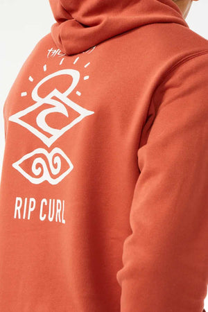 A person wears a rust-colored Rip Curl Search Icon Hood featuring white graphic designs and "RIP CURL" text on the back. Made from organic cotton, it showcases abstract shapes and patterns above the brand name.
