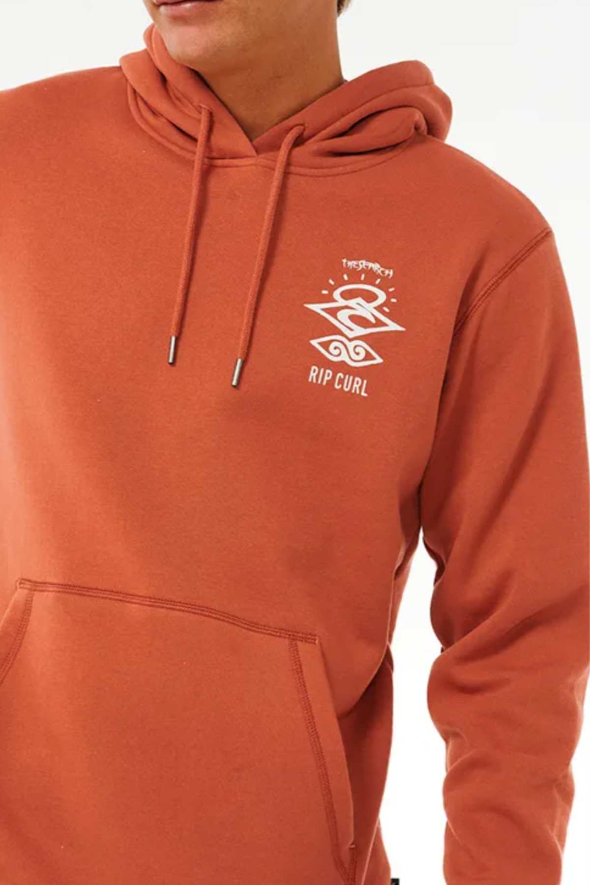 Someone wearing the Rip Curl Search Icon Hood—a rust-colored organic cotton hoodie with a white graphic logo and text on the chest, complete with a kangaroo pocket and drawstrings.