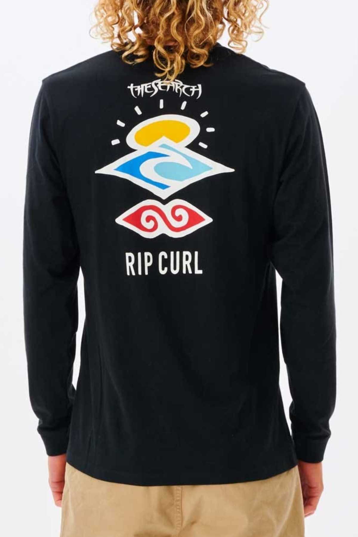 A person with curly hair wears the Rip Curl Search Icon Long Sleeve Tee—crafted from organic cotton jersey with colorful abstract graphics, "THE SEARCH" above and "RIP CURL" below on the back—paired with light-colored shorts.