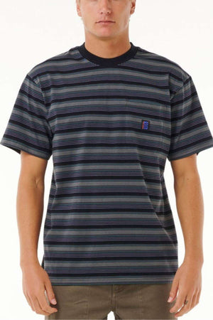 A person wearing the Rip Curl Search Stripe Tee, a black and gray top made from soft cotton, featuring a small embroidered patch on the chest, paired with olive green pants against a plain white background.