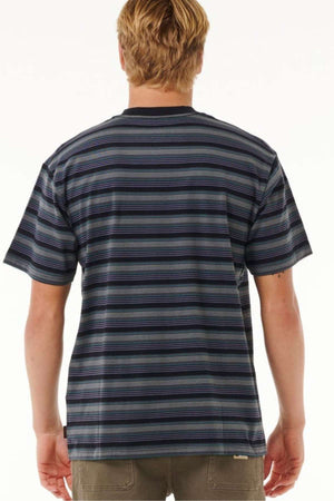 A person with blonde hair is seen from behind wearing a soft, dark Rip Curl Search Stripe Tee featuring horizontal stripes and beige pants. The background is plain white.