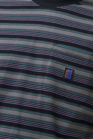 A detailed view of the Rip Curl Search Stripe Tee highlights its cotton fabric adorned with horizontal stripes in varying shades of blue, gray, and black. The left side sports a small pocket with a distinctive logo displaying an "S" design in orange and blue.