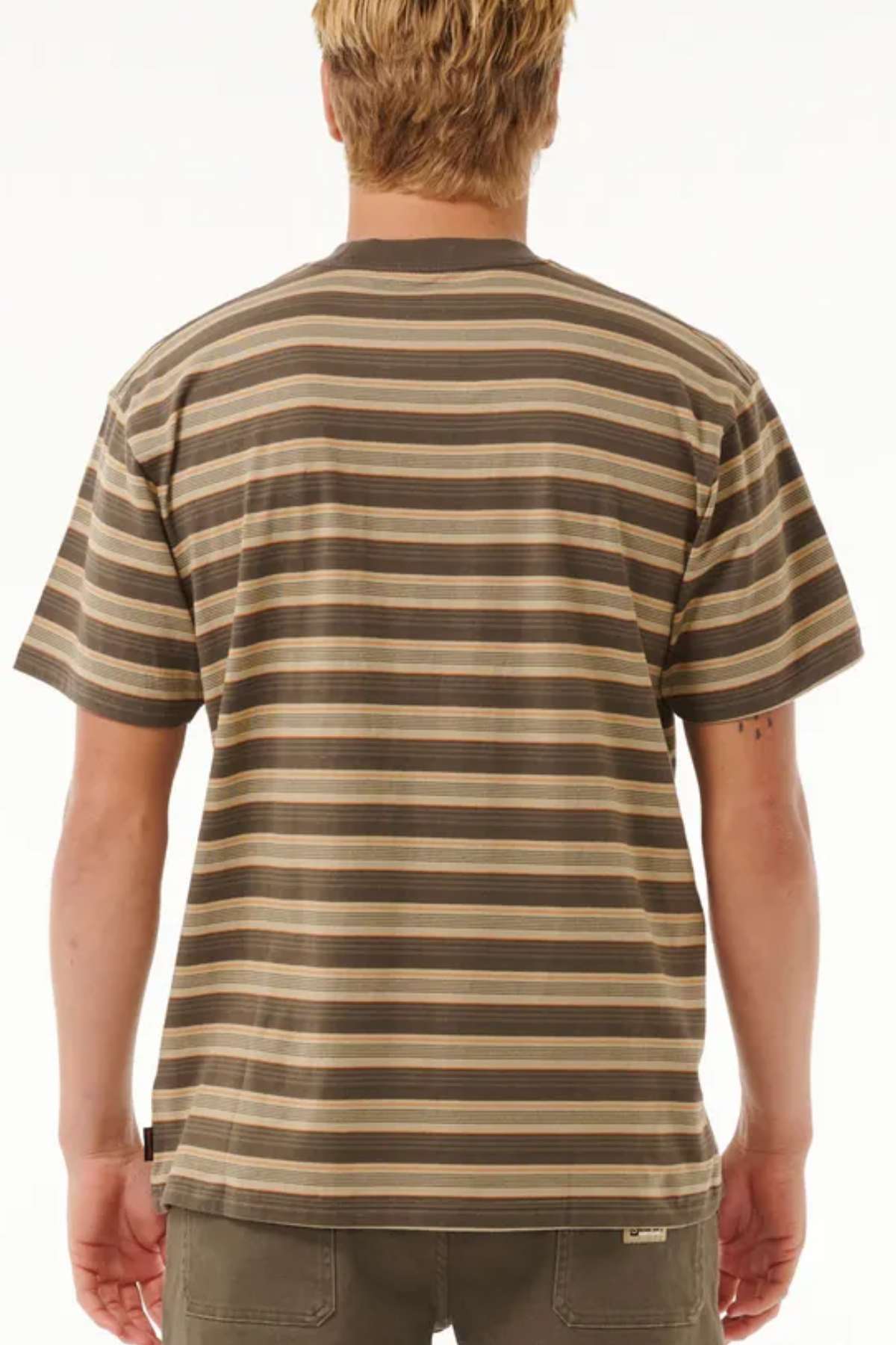 A person with short blond hair is wearing a Rip Curl Search Stripe Tee in brown and beige stripes, made from 100% cotton, facing away from the camera. They are also in dark pants against a plain white background.