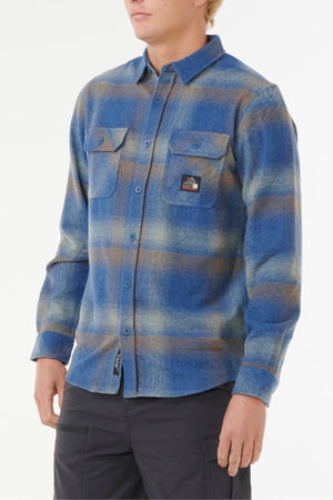 A person wears the Rip Curl Search Wool Flannel—a blue and tan plaid button-up with two chest pockets (one with a small patch), long sleeves, and dark pants. The background is plain white.