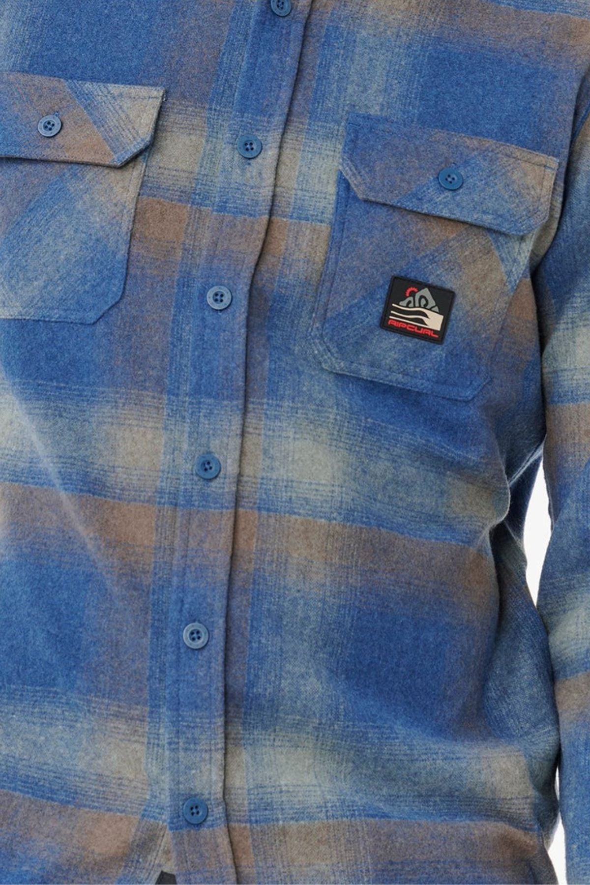A close-up of a person wearing the Rip Curl Search Wool Flannel—a blue and beige plaid wool button-up shirt with two chest pockets, one featuring a rectangular patch with a mountain and sun design—perfect for coastal living.