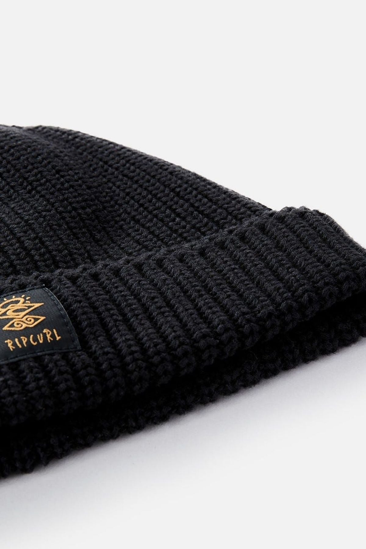 A close-up of the black Rip Curl Searchers Regular Beanie with a folded brim, showcasing a black and gold logo tag on the edge and surf-inspired style.