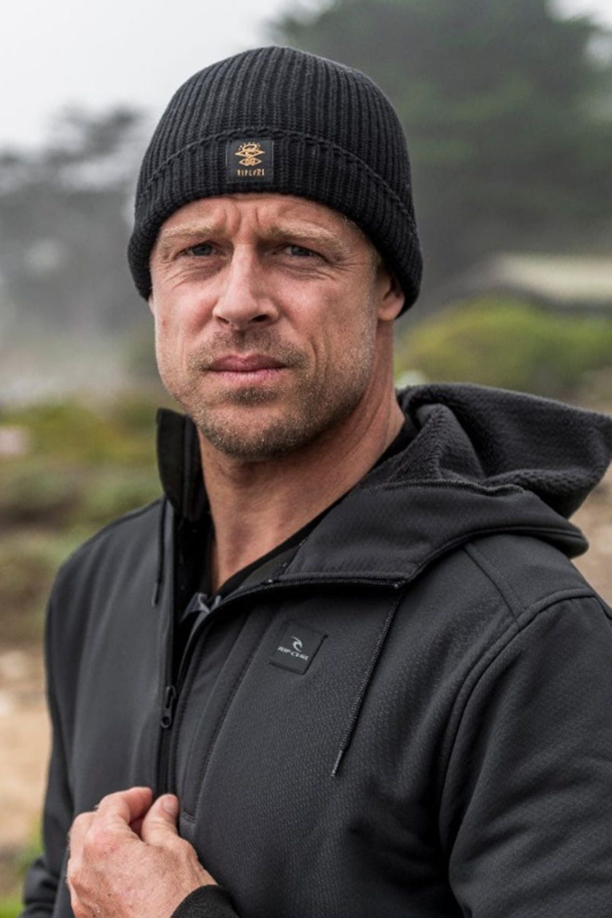 A person wears a black, surf-inspired Rip Curl Searchers Regular Beanie and a dark jacket with sherpa lining outdoors. The slightly blurred background shows greenery beneath a cloudy sky. They face the camera with a neutral expression.