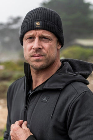 A person wears a black, surf-inspired Rip Curl Searchers Regular Beanie and a dark jacket with sherpa lining outdoors. The slightly blurred background shows greenery beneath a cloudy sky. They face the camera with a neutral expression.