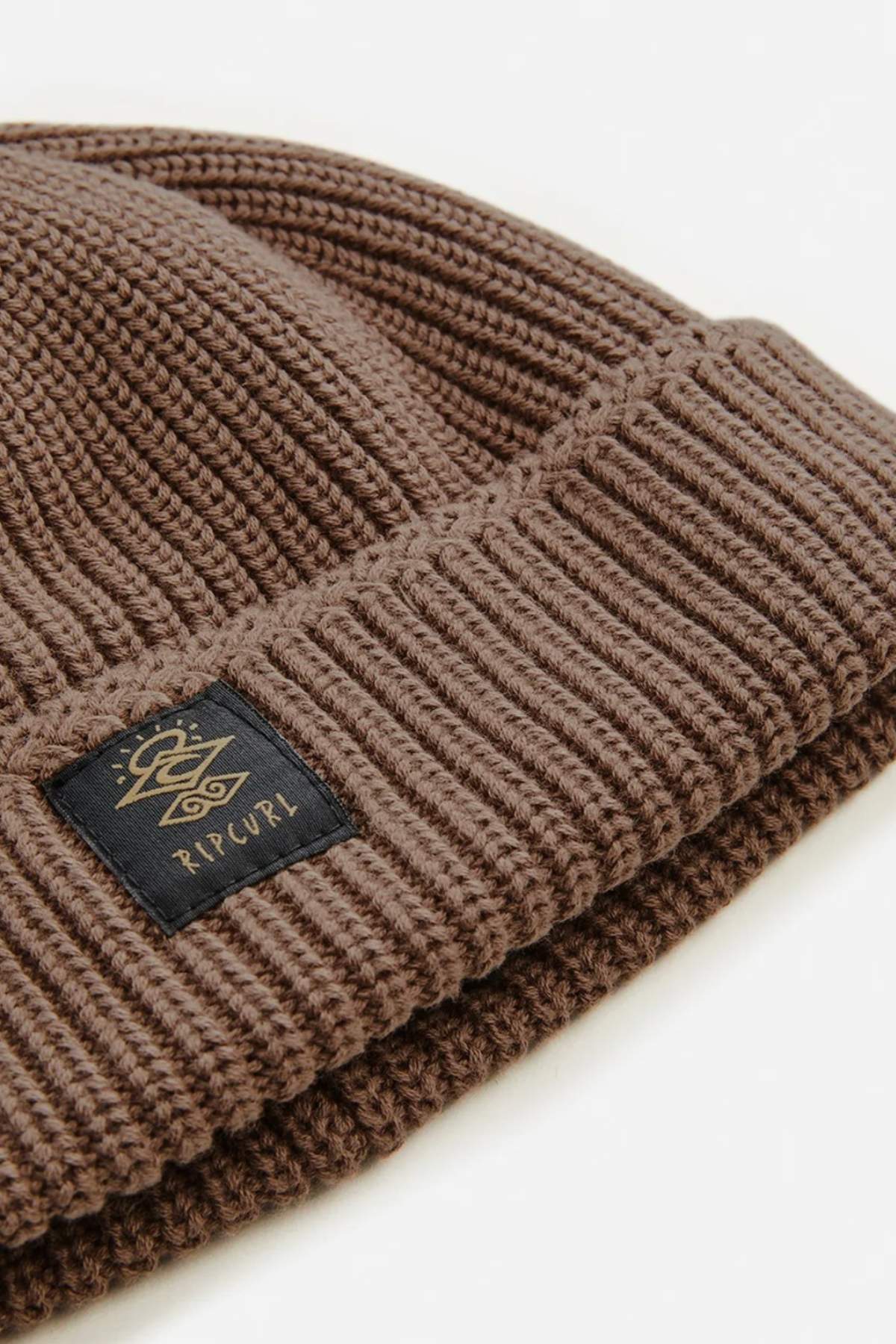 A close-up of the Rip Curl Searchers Regular Beanie highlights its cozy sherpa lining, folded cuff, and a black fabric tag with the "RIP CURL Searchers" logo in gold lettering.
