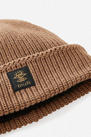 Close-up of the Rip Curl Searchers Regular Beanie in brown, featuring a folded brim and a black patch with the gold Rip Curl logo and text for surf-inspired style.