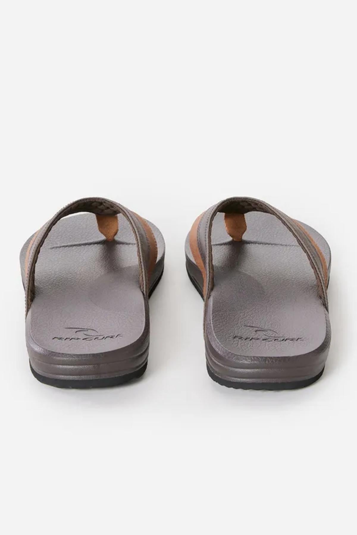 The Rip Curl Soft Top Thongs are showcased from the back, slightly elevated to highlight their raised soles and the textured footbed with a Rip Curl logo embossed on each sole. The flip-flops feature wide, padded straps for added comfort against a clean white background.