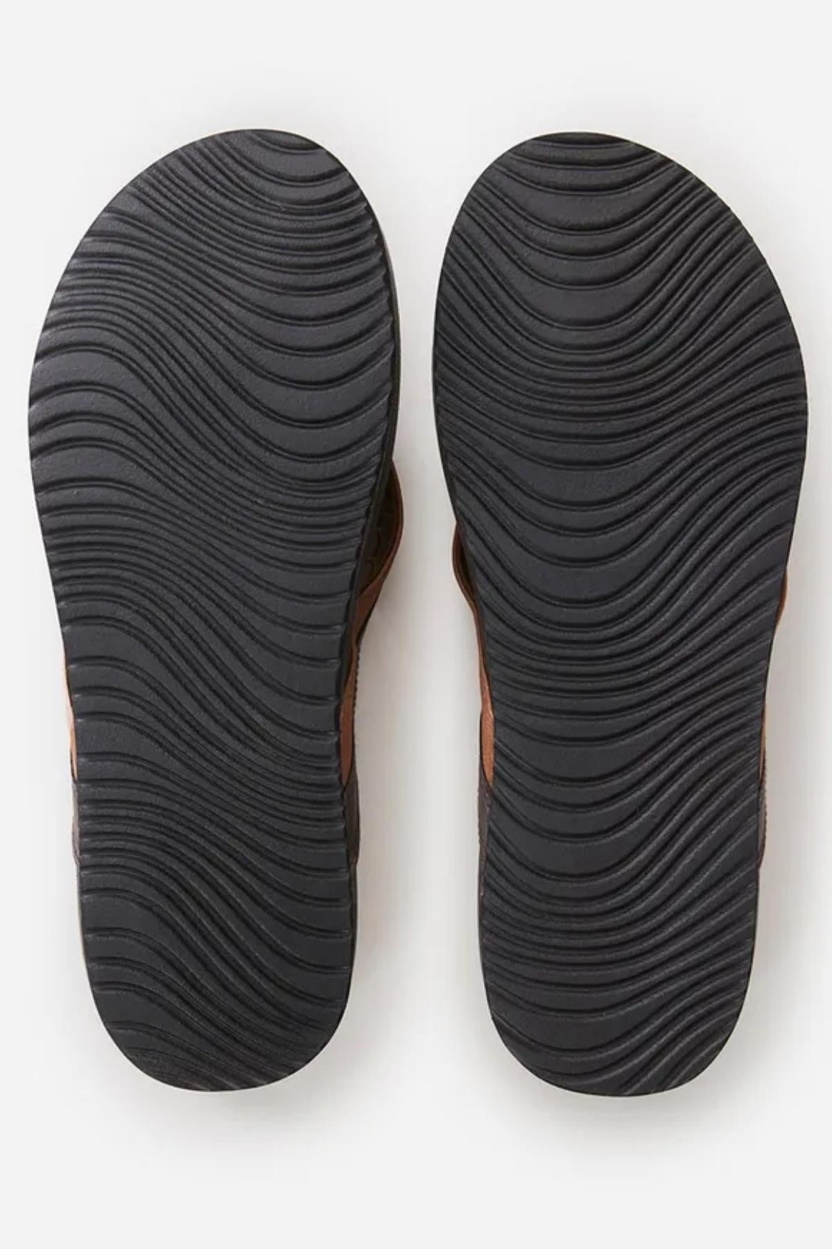 A close-up image of the soles of a pair of Rip Curl Soft Top Thongs. The soles are black with a wavy, textured pattern providing traction. The thongs themselves are brown, visible slightly along the edges of the image. The background is plain white.