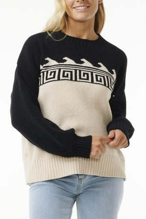 A woman stands against a white background wearing the Rip Curl Soleil Knit Crew, an oversized recycled poly chenille sweater with geometric wave and Greek key patterns, paired with light blue jeans.