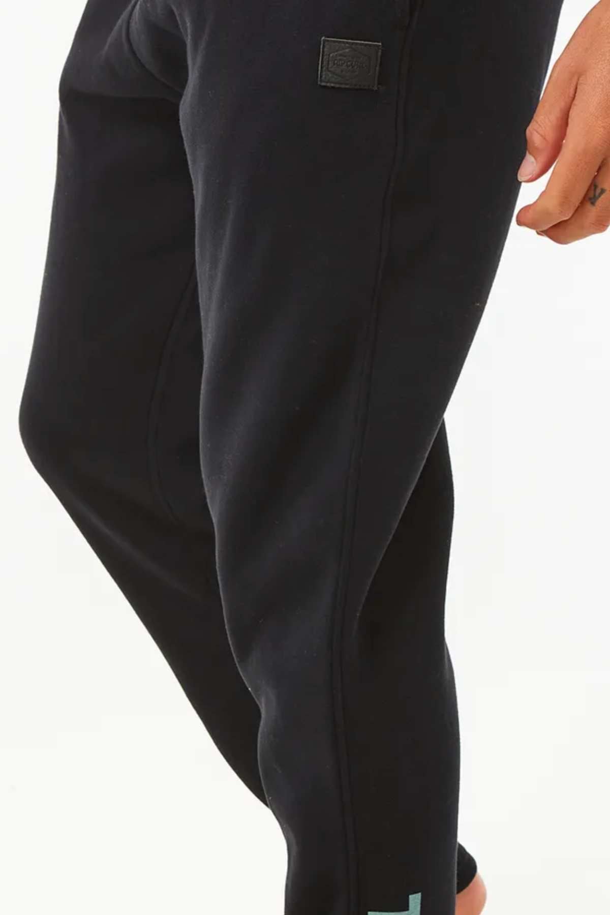 A person shown from the waist down wears Rip Curl Stapler Trackpant joggers with an elastic waistband; part of their hand is visible on the right side, set against a plain white background.