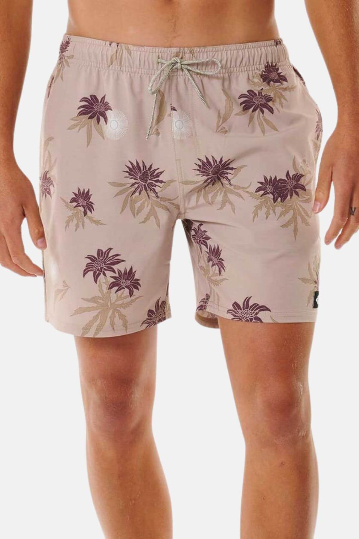 The Rip Curl Sun Razed Floral Volley shorts feature a light brown floral pattern in purple and beige. Made from quick-dry fabric, they include a drawstring waist and two small reflective circles on the front. A plain white backdrop highlights their style and performance.