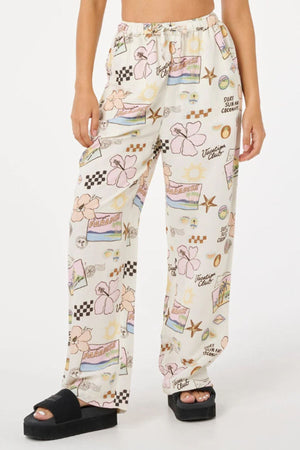 A person wears Rip Curl Sunset Luau Wide Leg Pants in a linen-viscose blend with vibrant summer prints, styled with black platform sandals. The warm-weather look pops against the plain white background.