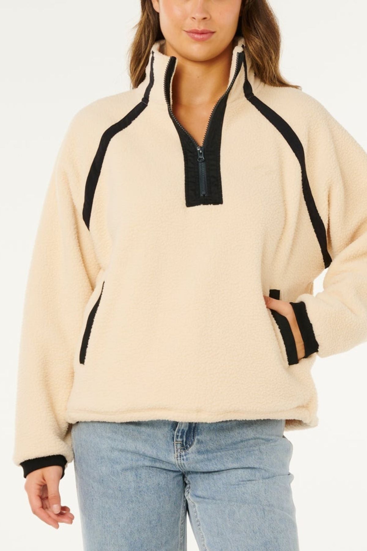Wearing the Rip Curl Surf Club Heritage Polar fleece pullover in cream with black trim and a half-zip front, paired with light blue jeans, a person poses with one hand in their pocket against a white background.